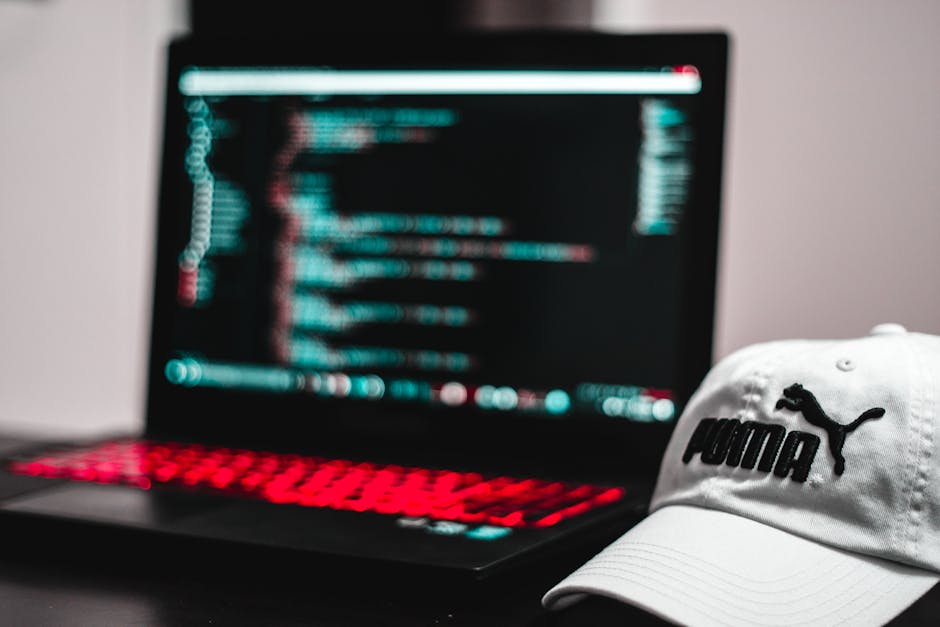 A close-up view of a modern laptop with a blurred screen displaying lines of code or digital data, placed on a dark surface. Next to the laptop, a white baseball cap branded with the Puma logo rests c